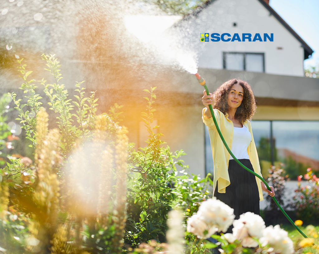 A happy woman in a yellow cardigan outside watering her garden with a long green water hose